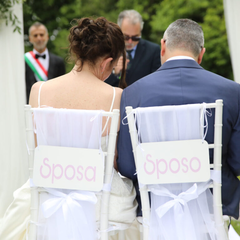 Sposi di schiena seduti su sedie decorate con cartelli in alluminio decorativi con scritto sposa e sposo